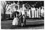 Children pose with woman dressed in antebellum gown in front of antebellum style home by Edwin E. Meek