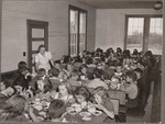Lone worker serving approximately 50 children in a cafeteria filled with trestle tables. by USDA